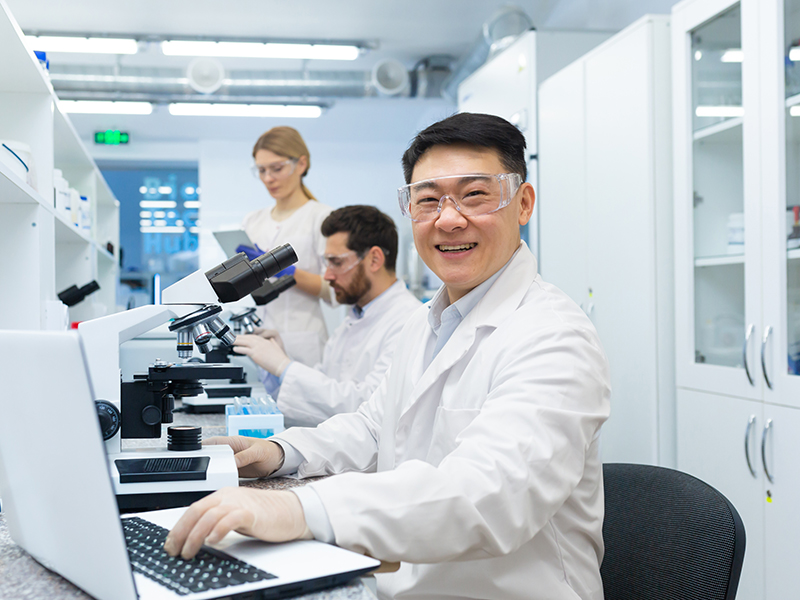 Portrait of asian lab technician, man with team group of people working in laboratory researching, man with microscope in white coat and goggles smiling and looking at camera.